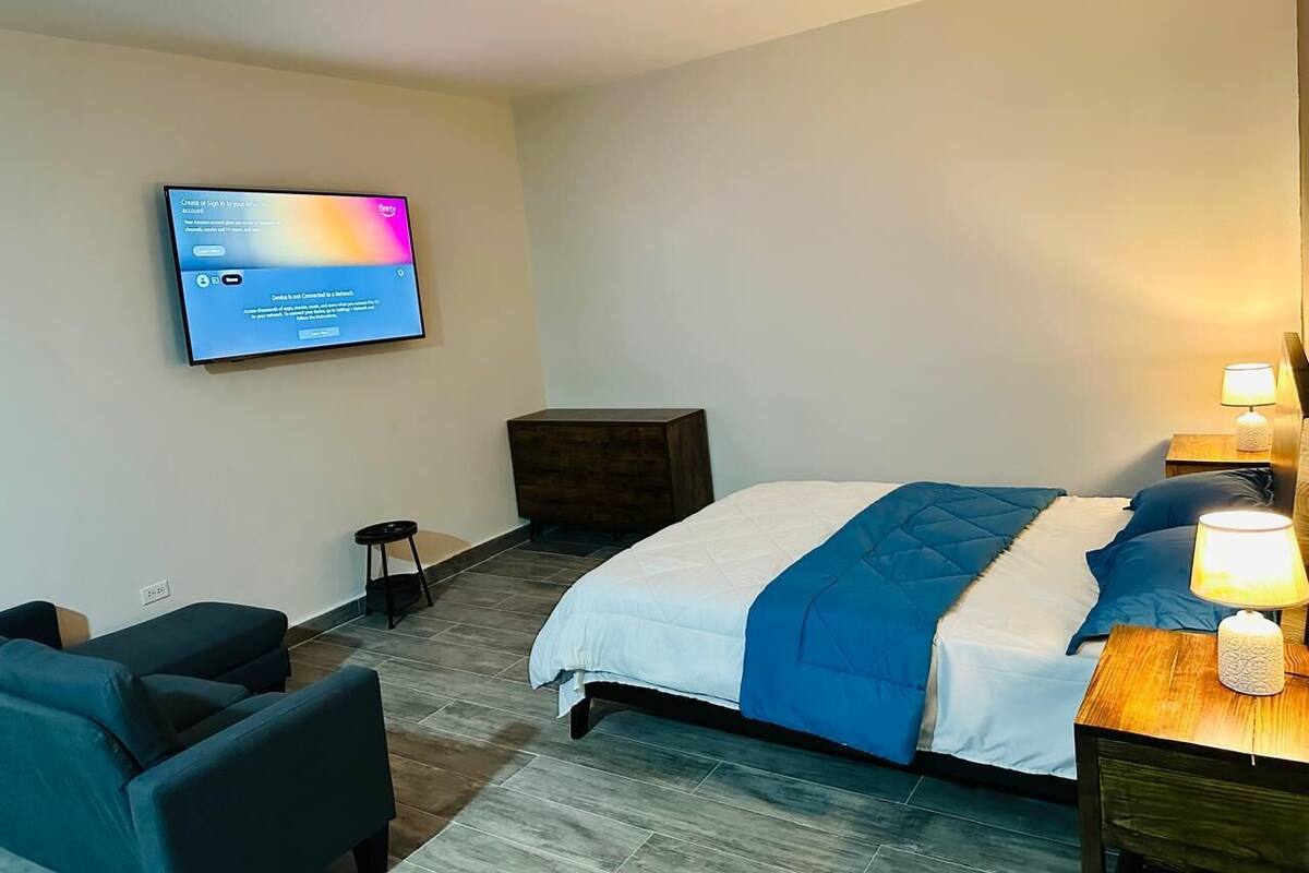 A hotel room with a bed, a desk with a lamp, a TV mounted on the wall, a small stool, and a dark wooden dresser.