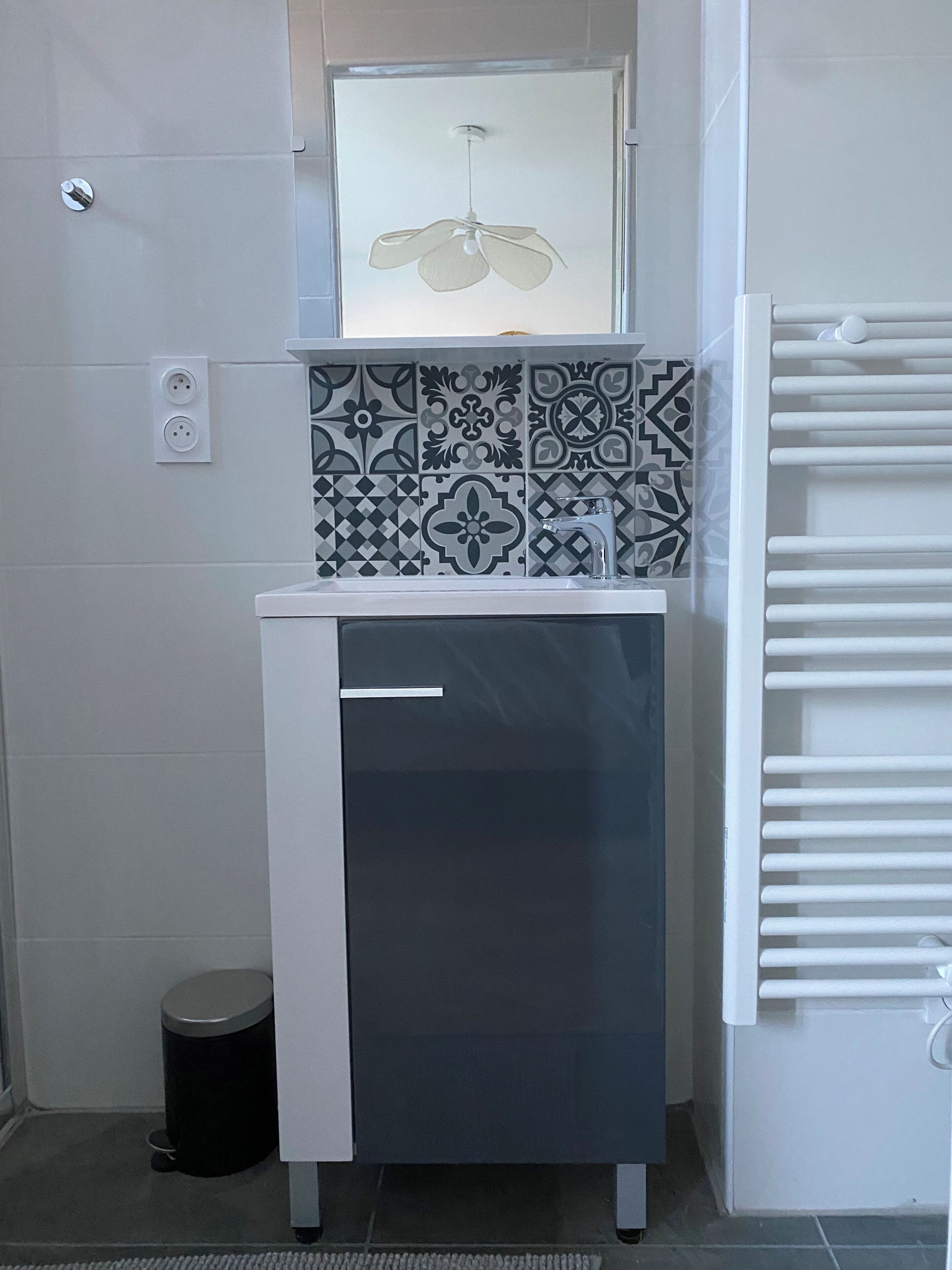 Une petite salle de bain avec un meuble blanc, une poubelle noire, un pare-chocs en carreaux décoratifs et un ventilateur de plafond.