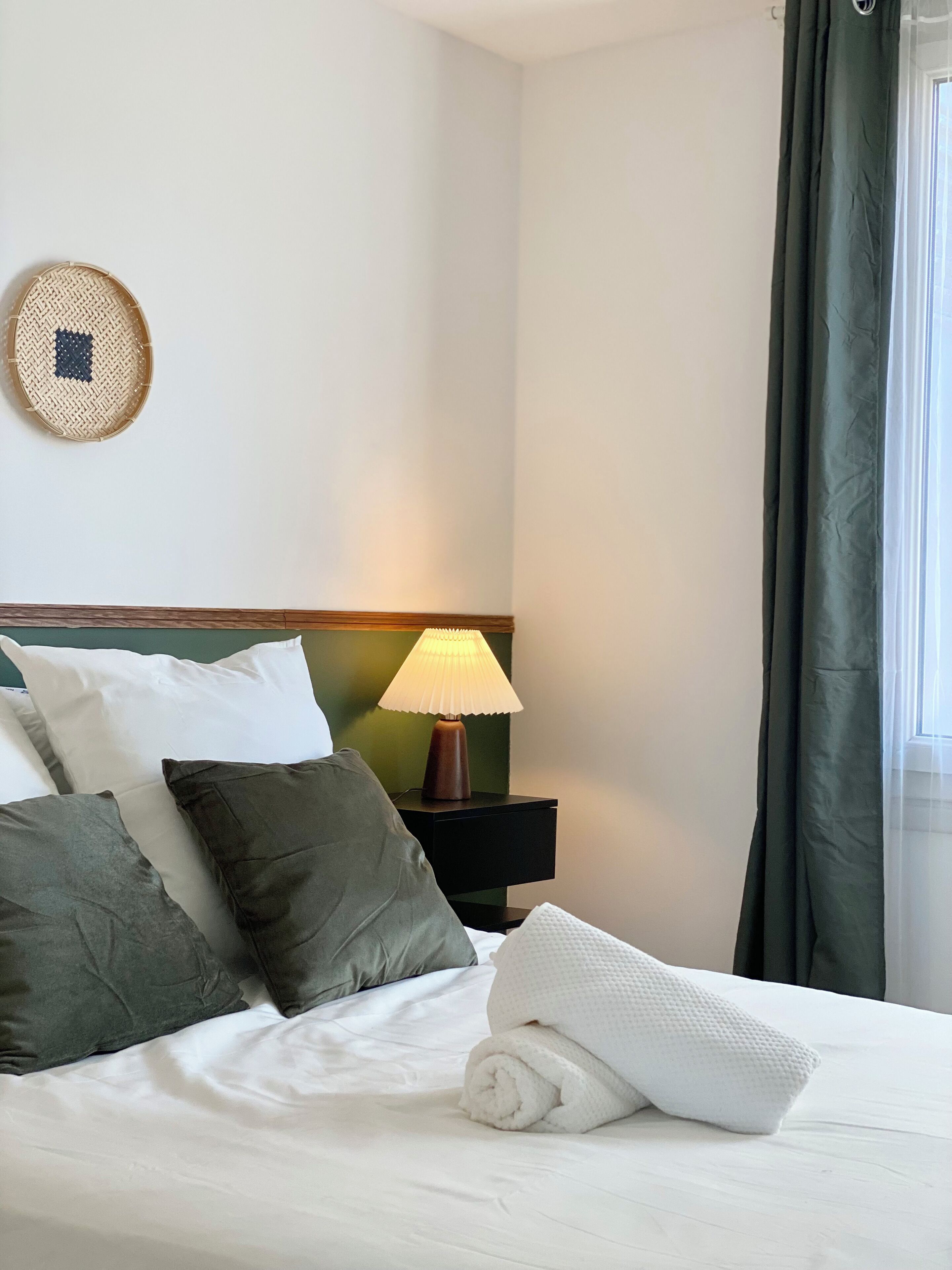 A neatly made bed with pillows and a rolled towel, a bedside lamp, and a wall-mounted decorative item.