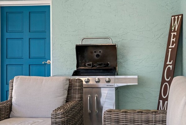 Una habitación con puerta azul, sillas de mimbre, una pequeña cocina americana con una parrilla y un cartel que dice 'WELCOME'.