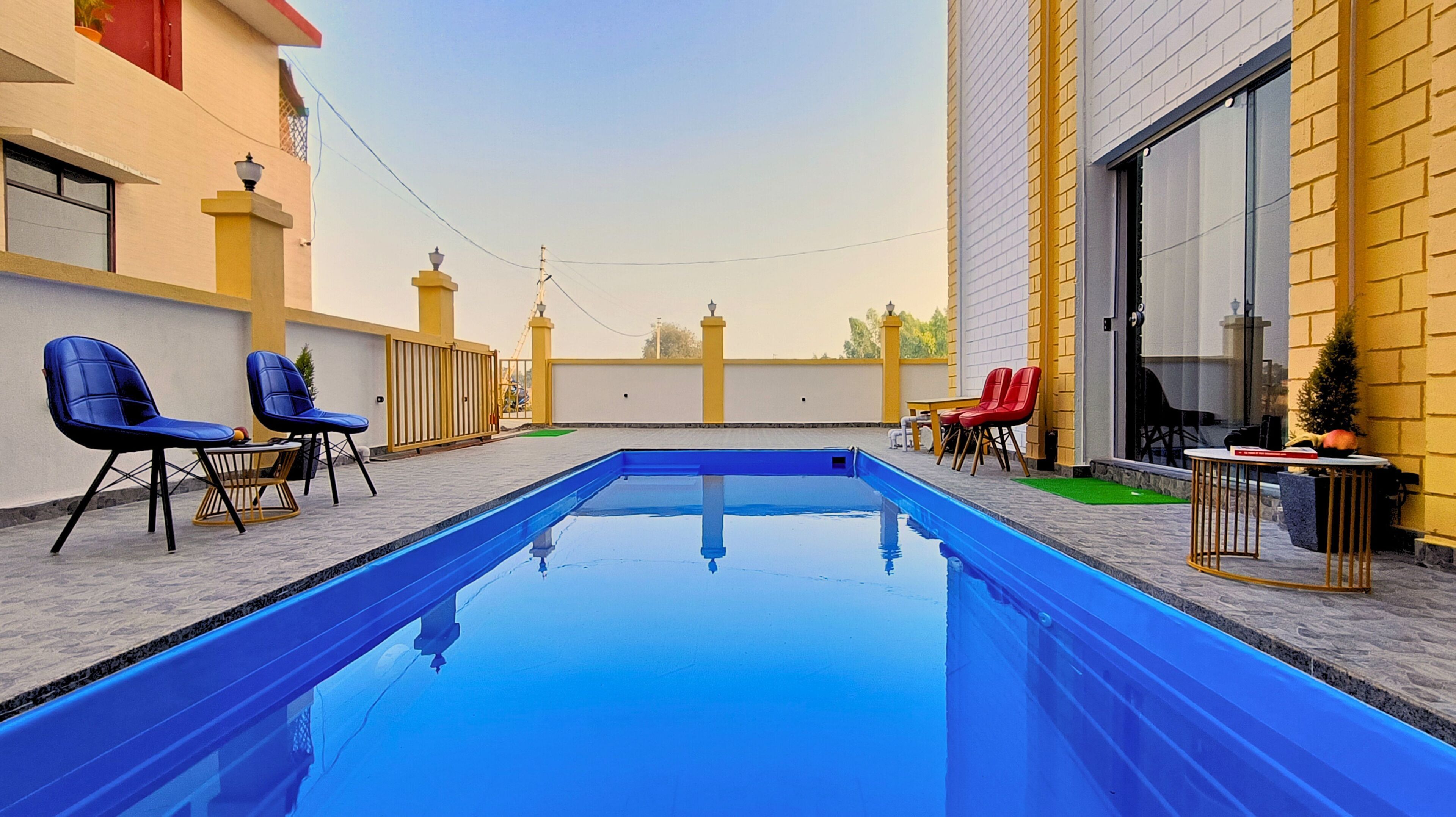 Una piscina con agua azul, rodeada de mobiliario exterior con sillas azules y rojas, y un edificio amarillo con un ventanal grande.