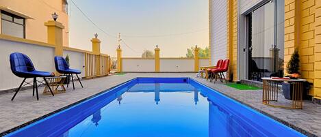 Una piscina con agua azul, rodeada de mobiliario exterior con sillas azules y rojas, y un edificio amarillo con un ventanal grande.