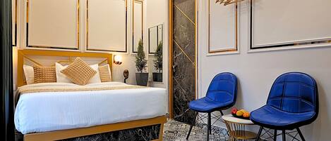 A hotel room with a large bed, two blue chairs, a small table, and a decorative mirror.
