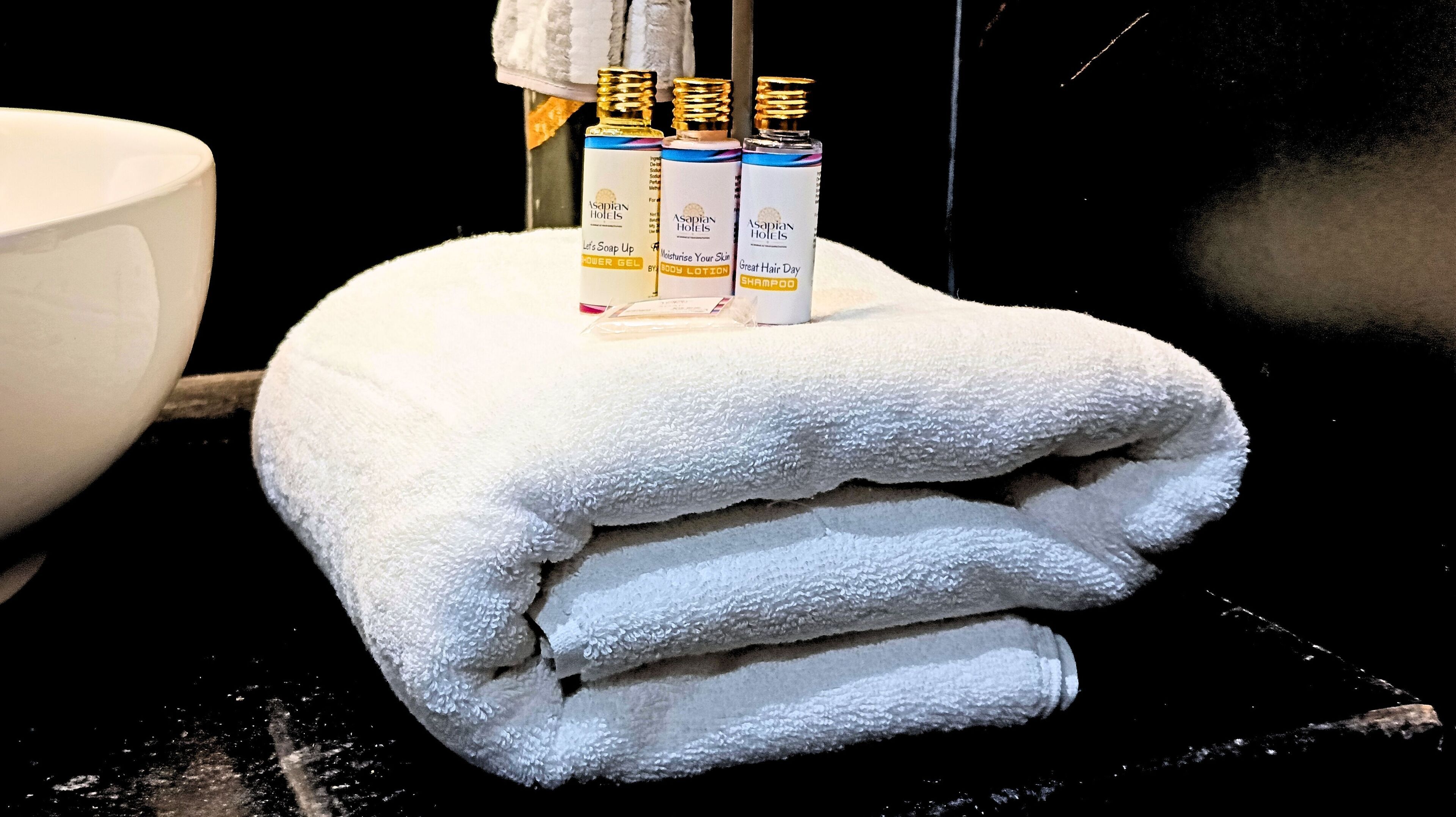 Een stapel witte handdoeken met drie flessen Asapian Hotels producten erboven.