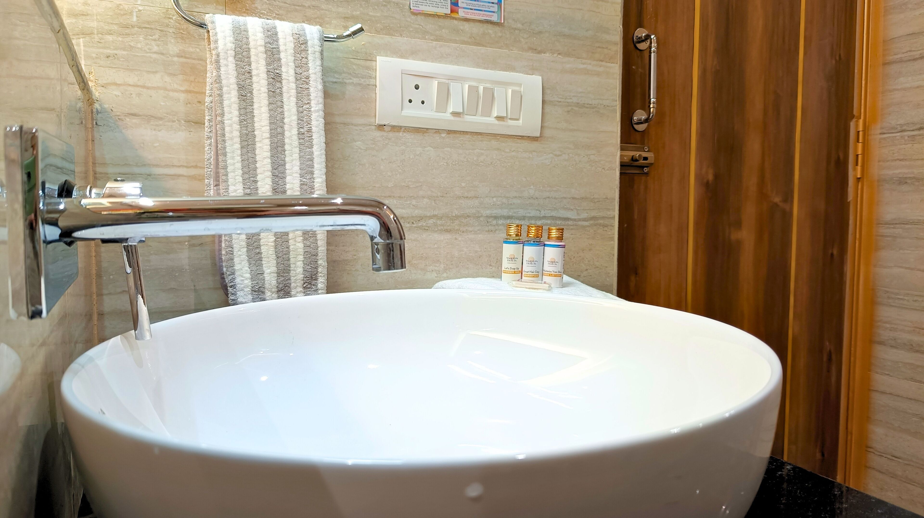 A bathroom with a large white sink, a towel hanging on a rack, a faucet, a wooden door, and a wall socket.