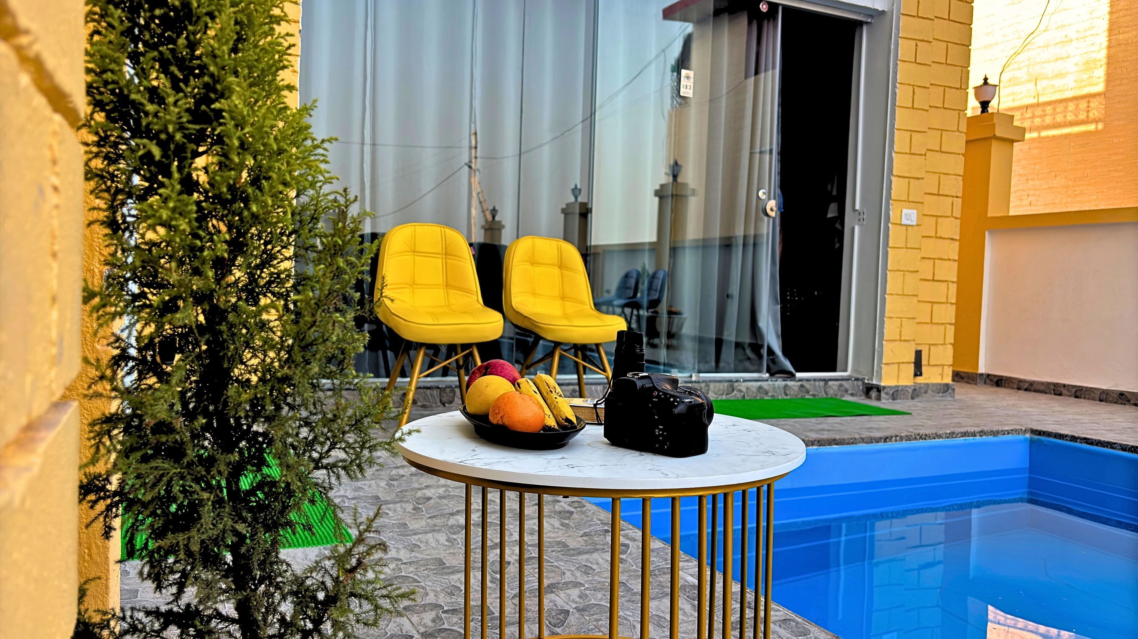 Een zwembadterras met een marmeren tafel, twee gele stoelen, een schaal fruit en een camera.