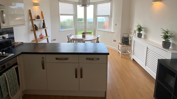 A modern kitchen with white cabinets, a black countertop, and a dining area with a table and chairs. There are windows with blinds, a hanging light fixture, and a radiator with plants.