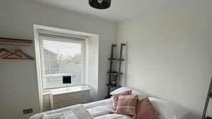A neatly arranged bedroom with a bed, bedside tables, a window with blinds, a hanging light fixture, and a shelf unit.
