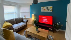 A living room with a brown leather sofa, a grey sofa with yellow cushions, a wooden coffee table, and a flat-screen TV on a stand.