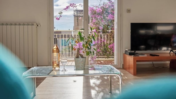 A living room with a glass table, a bottle, a wine glass, a TV, and a balcony with pink flowers.