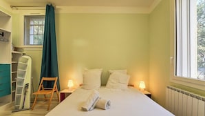 A bedroom with a bed, two bedside lamps, a window with a curtain, a wooden chair, and a mirror on the wall.