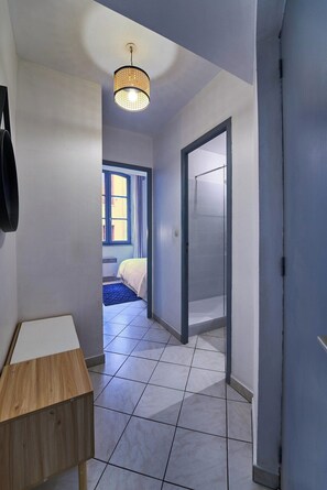 Un couloir avec une table en bois, un luminaire suspendu et une porte donnant sur une chambre à coucher équipée d’un lit et d’une fenêtre.