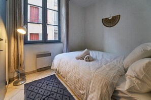 Et soveværelse med seng, radiator, et vindue med gardiner, en gulvlampe og en vægmonteret ventilator.