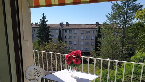 Un balcón con una mesa y sillas blancas, un jarrón con flores y vistas a edificios y árboles.