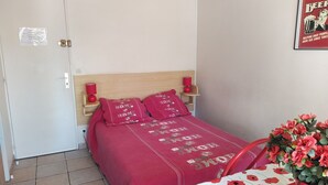 Un dormitorio con cabecera de madera, una cama con manta roja, una mesita roja con una maceta y un cartel en la pared.