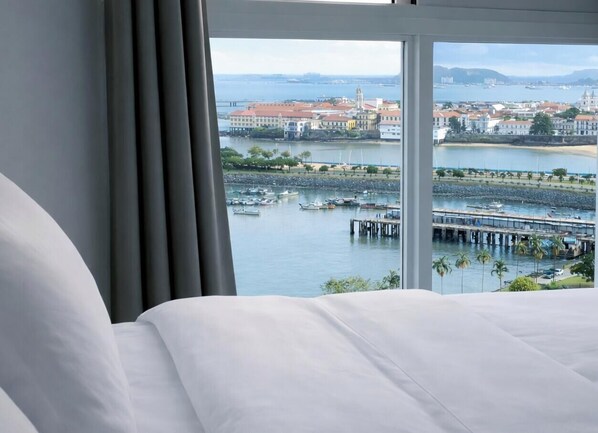 A hotel room with a large bed, a view of a waterfront with boats and a pier, and a cityscape in the background. - 80m² Luxury Ocean View Apartment PH The Sands, Next to Hilton (Panama City)
