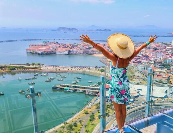 A person standing on a high vantage point overlooking a coastal city with a bridge, boats, and buildings. - 80m² Luxury Ocean View Apartment PH The Sands, Next to Hilton (Panama City)