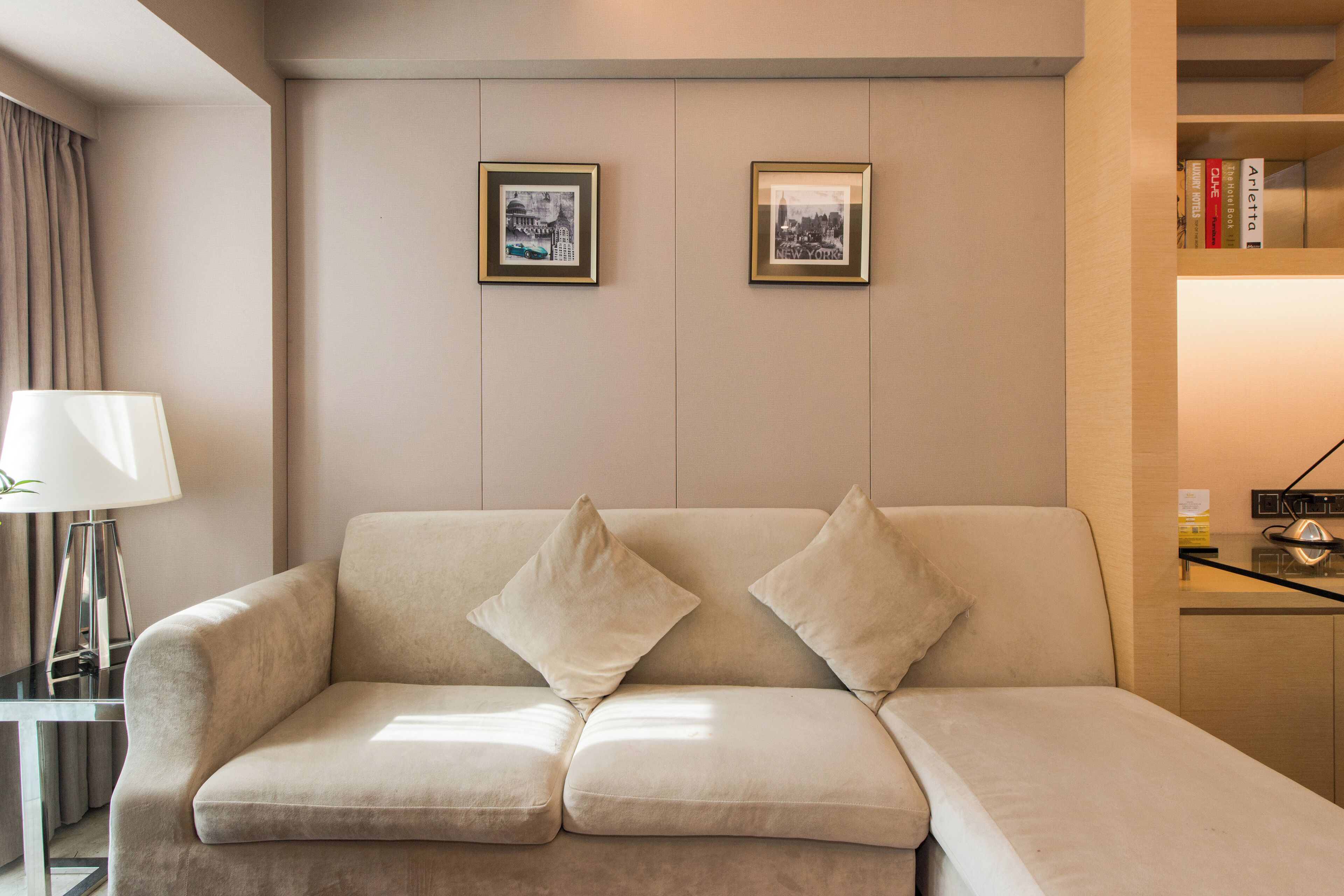 Ein modernes Wohnzimmer mit einem beigefarbenen Sofa, zwei gerahmten Bildern an der Wand, einer Lampe und einem Bücherregal.