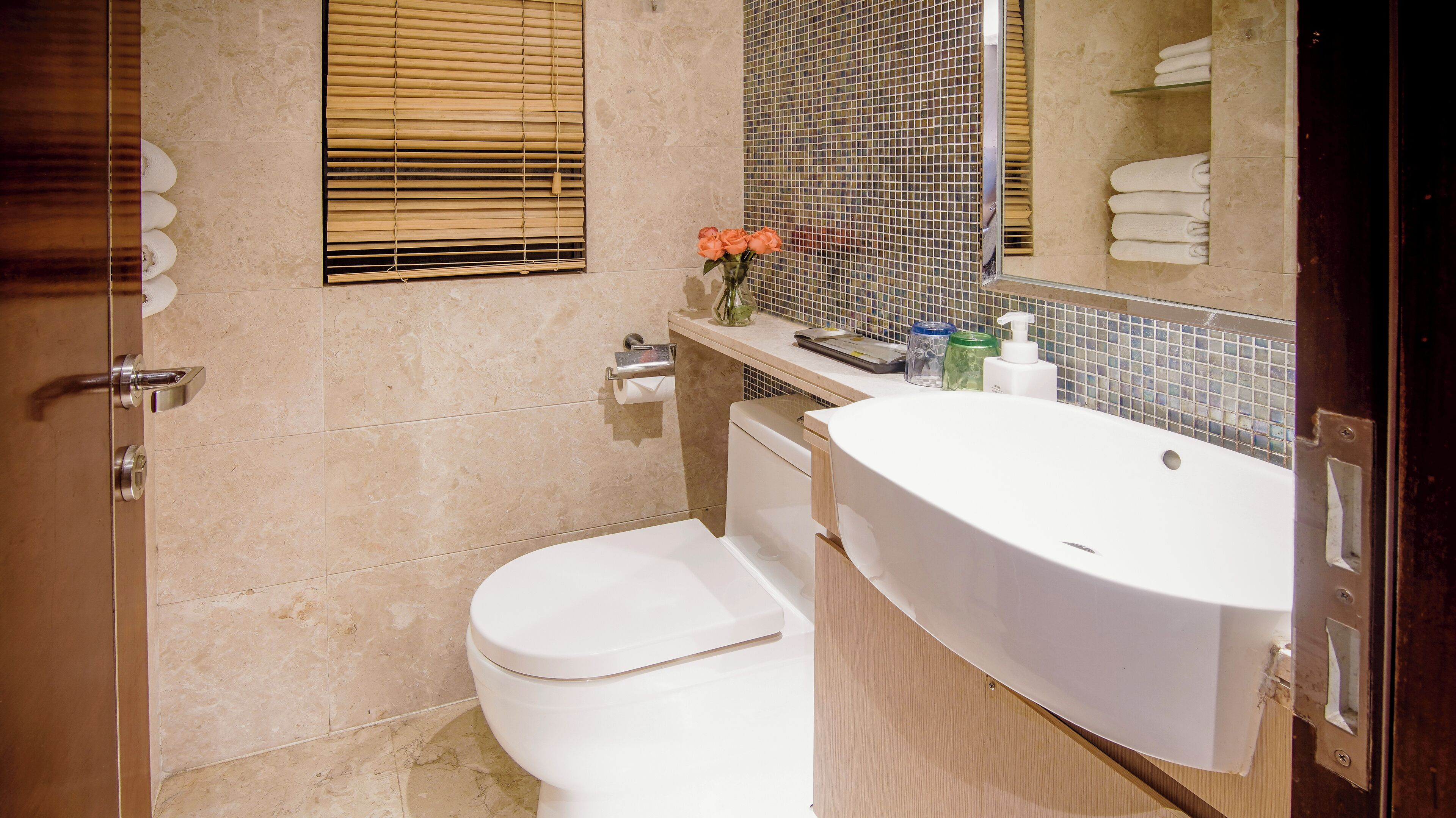 A bathroom with a toilet, sink, and mirror. There is a window with blinds and a towel rack with towels.