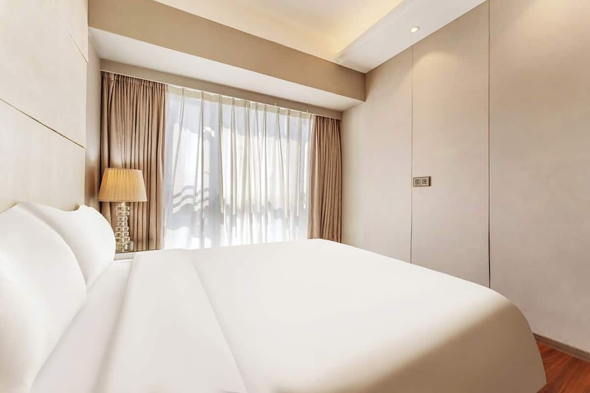 A modern hotel room with a large bed, a bedside table with a lamp, a window with sheer curtains, and a wardrobe.