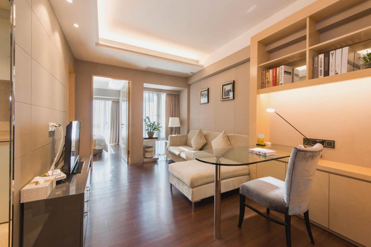 A modern hotel room with a glass table, a sofa, a TV, and a bookshelf.