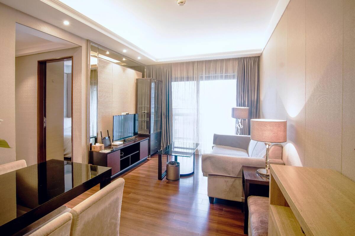 A modern hotel room with a large mirror, a flat-screen TV, a sofa, a small table, and a vase with flowers.