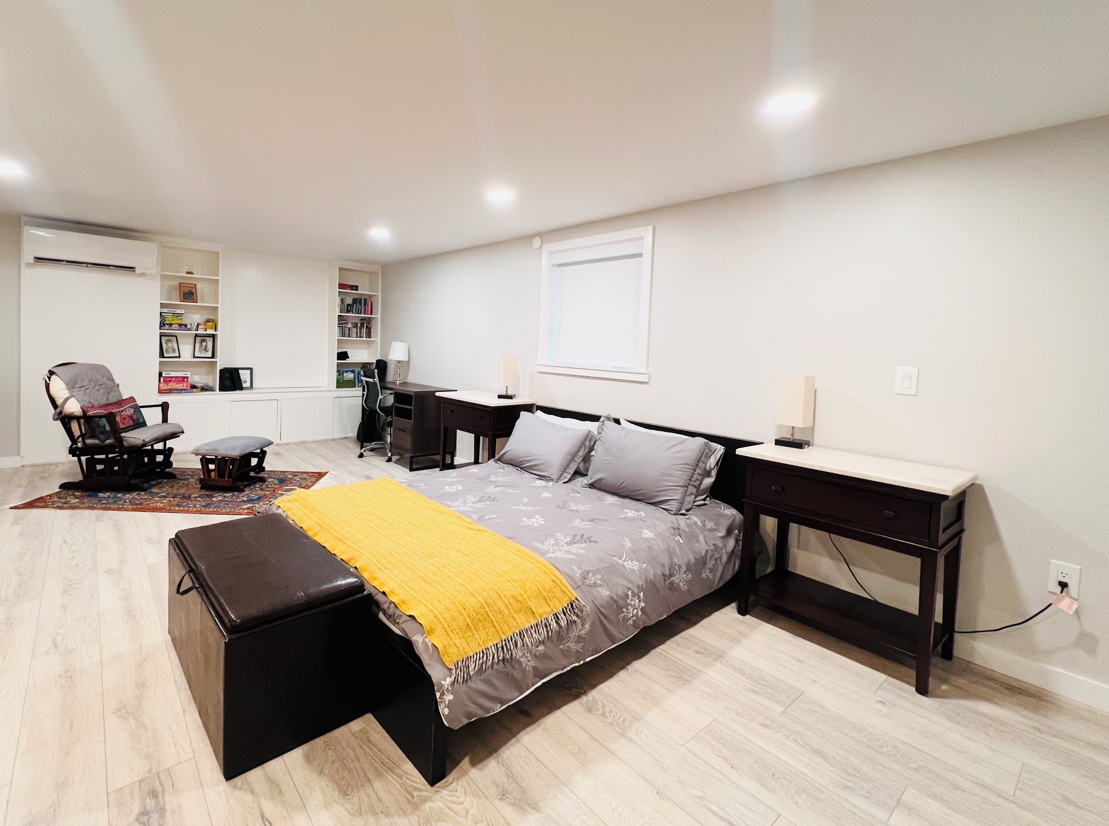 A bedroom with a bed, bedside tables, a desk, a chair, a bookshelf, and an air conditioning unit.
