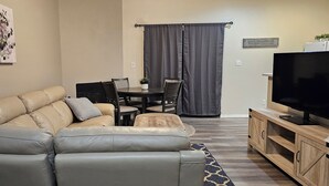 A living room with a beige leather sofa, a wooden coffee table, a TV on a wooden stand, and a dining area with a table and chairs. - Charming 3-bedroom apartment in enchanting Rio Rancho with AC, WiFi (Rio Rancho)