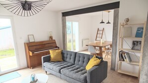 A living room with a grey sofa, a wooden piano, a dining table, and a bookshelf. - Large, bright, quiet house with piano and sunny enclosed garden. (Guilvinec)