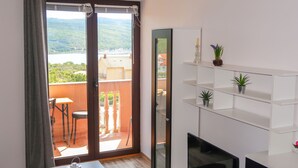 A modern living room with a TV, white shelving, and a balcony with a view of a lake and hills. - Apartment Rosić by Interhome (Krk/Krk)