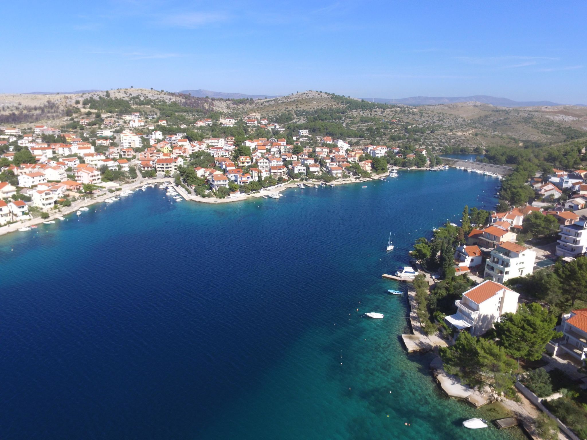 An aerial view of a coastal town with buildings along the shoreline, a clear blue sea, and hilly terrain in the background.