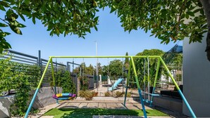 Un terrain de jeux avec des balançoires et un toboggan, entouré de verdure et sous un ciel dégagé.