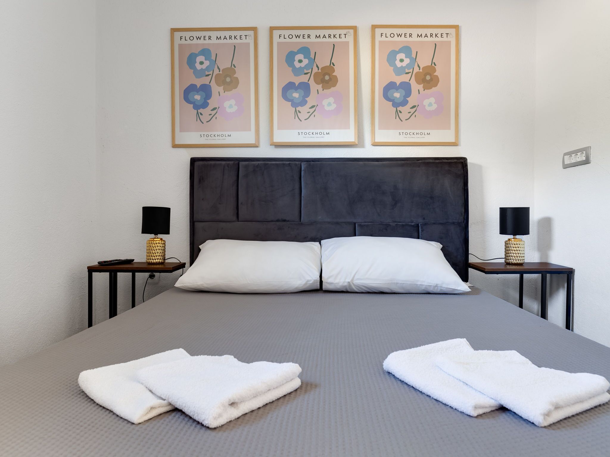 A neatly made bed with white pillows and two folded towels on the bed. There are three framed posters on the wall above the bed, each labeled "Flower Market Stockholm". The room has a simple, modern design with a grey headboard and two bedside tables with lamps.
