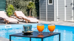 Une table placée au bord de la piscine, avec deux verres de jus d’orange et une tasse bleue, entourée de chaises longues et d’un abri de jardin en arrière-plan.
