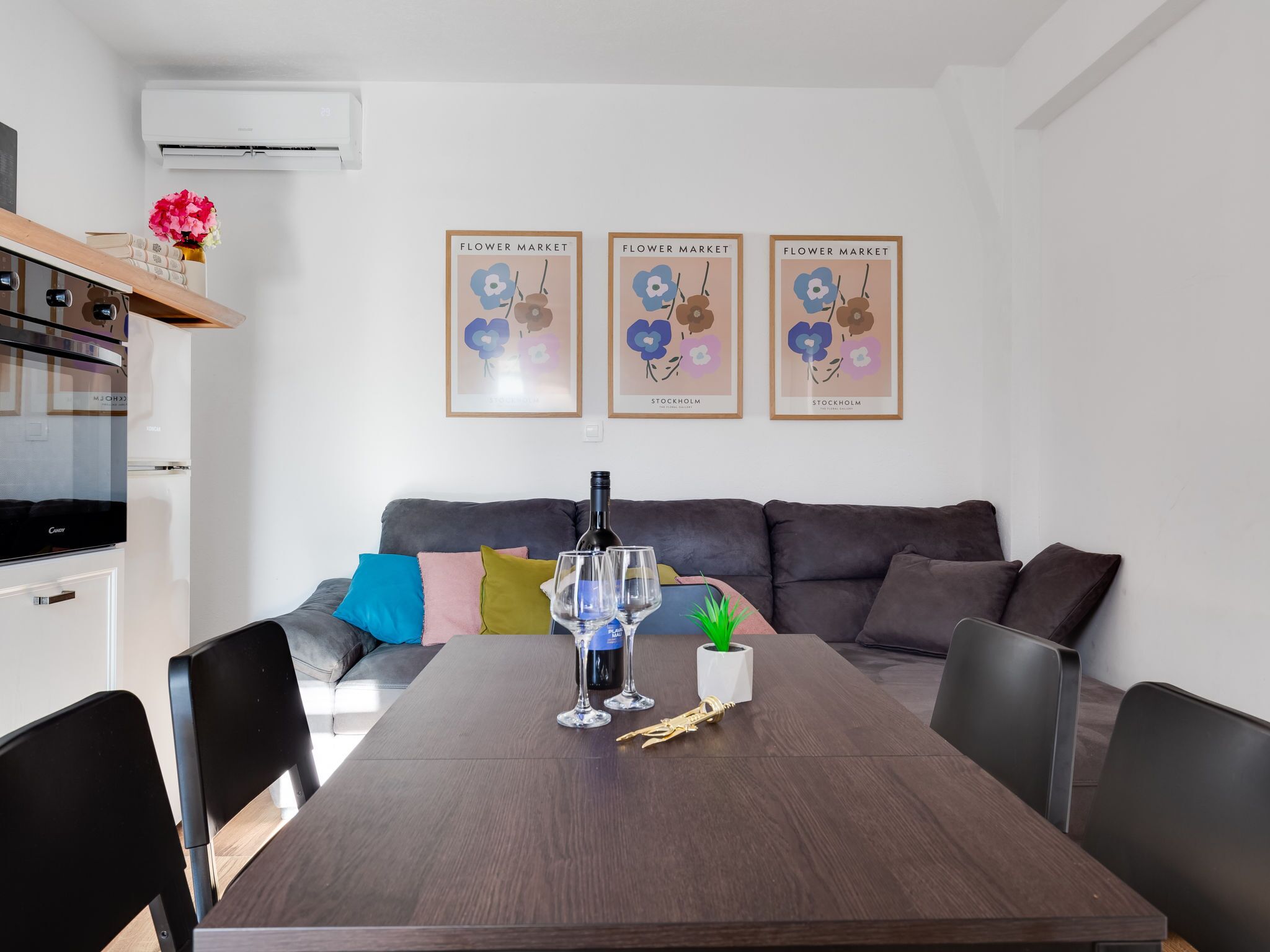 A modern living room with a dark wood dining table, a grey sofa with colorful cushions, and three framed posters on the wall.