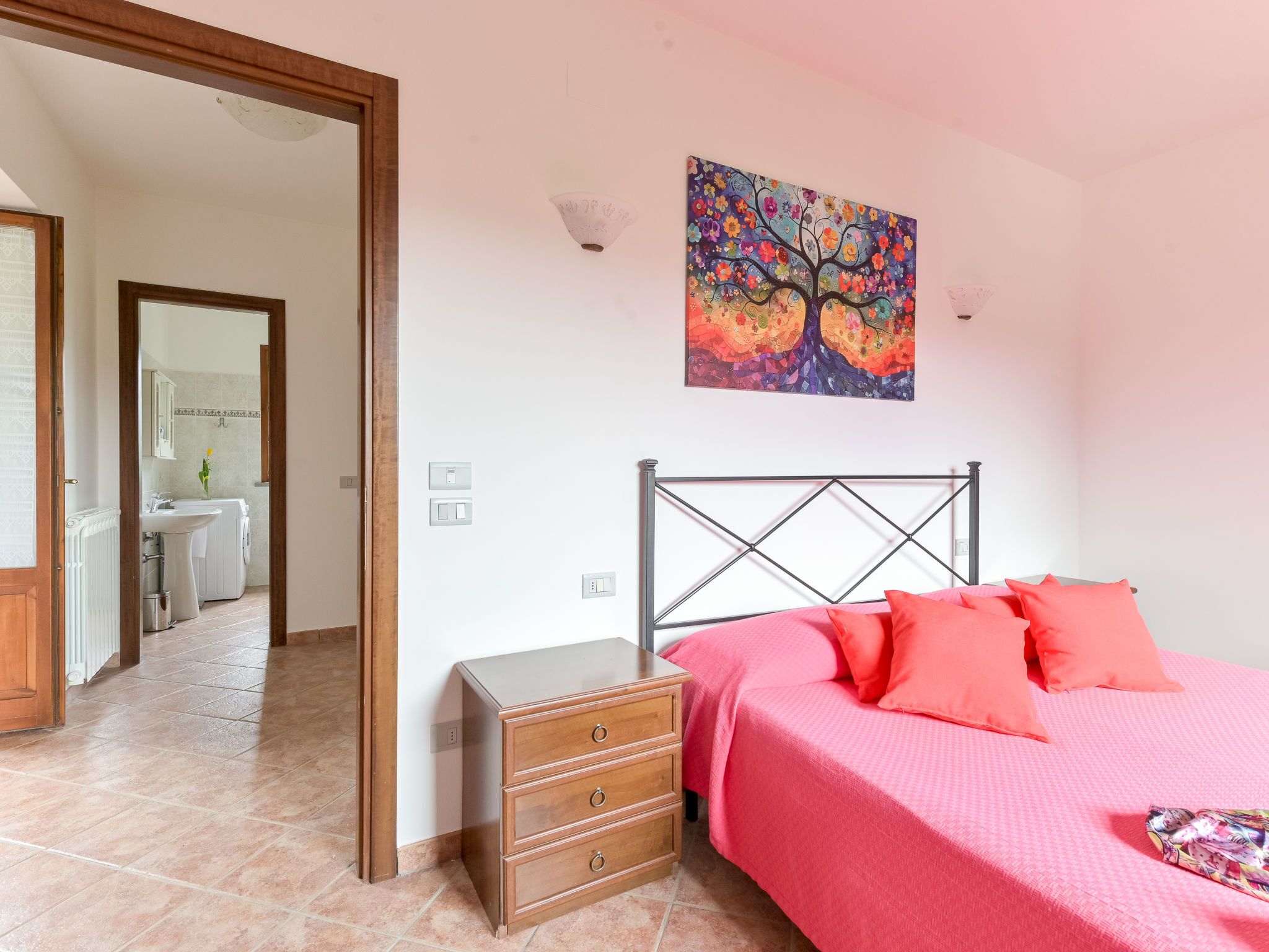 A bedroom with a bed, a nightstand, a dresser, a painting, and a bathroom visible through an open door.