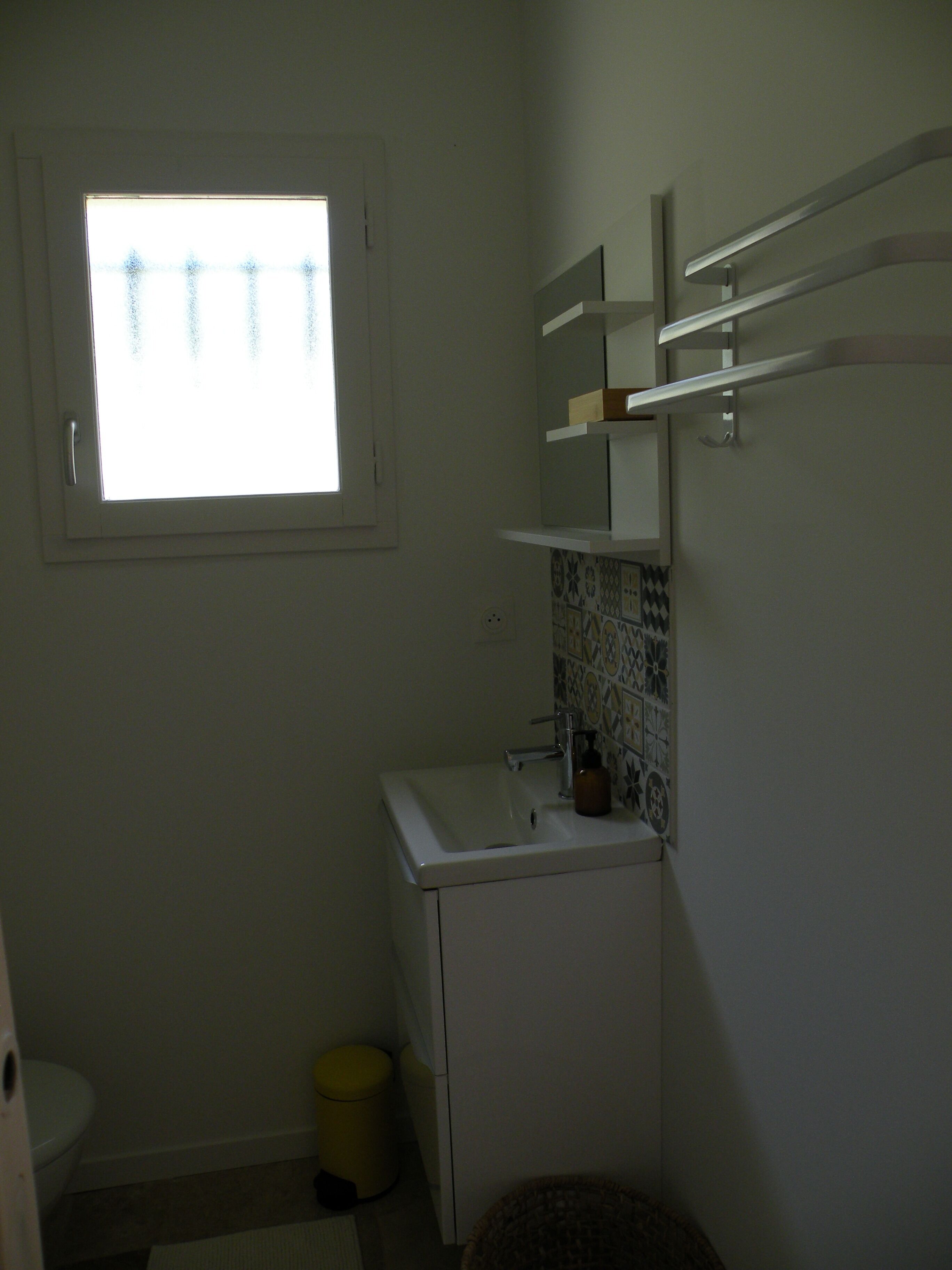 A small bathroom with a window, a sink, a toilet, and a trash bin.