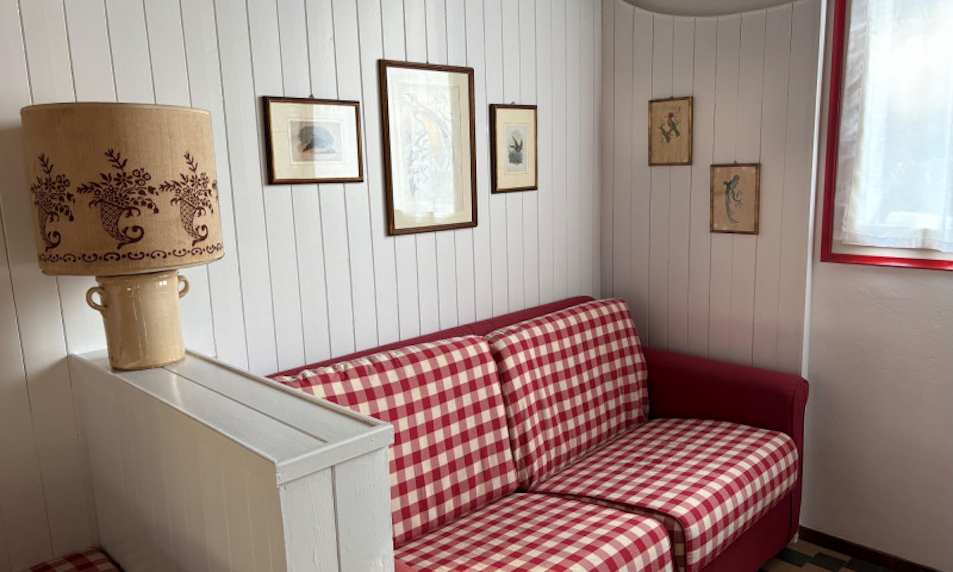 Une pièce avec un canapé à carreaux rouges et blancs, une lampe à motifs floraux et plusieurs tableaux encadrés accrochés au mur.