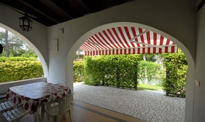An outdoor patio with a table and chairs, a red and white striped awning, and a view of neatly trimmed hedges. - Green oasis in Lignano Pineta (Lignano Sabbiadoro)