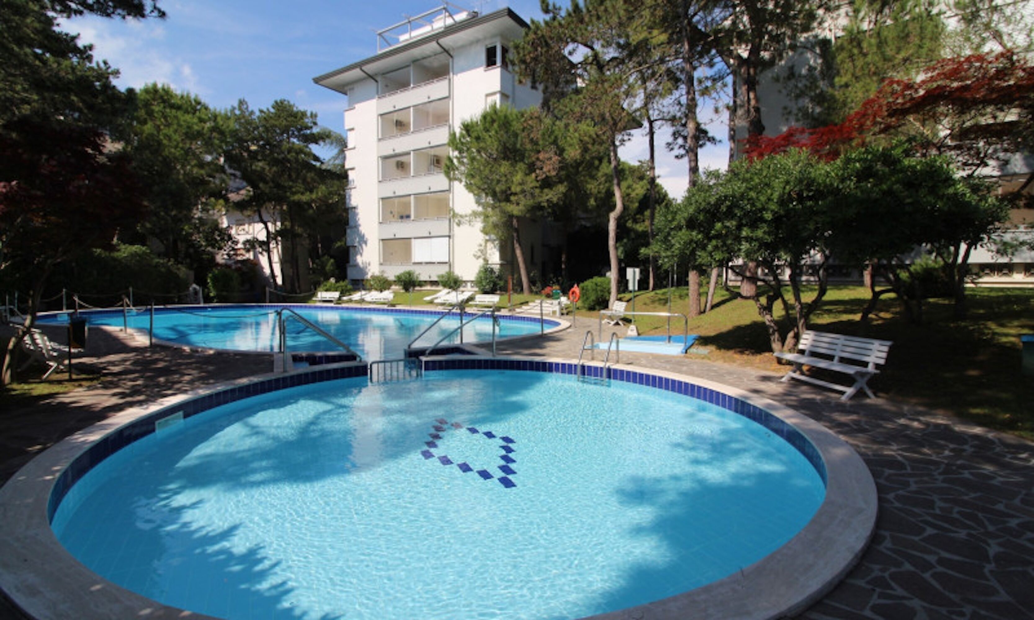 A circular pool with a blue center, surrounded by a stone deck, trees, and a white building in the background.