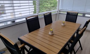 Un coin repas avec une table en bois et des chaises noires, de grandes fenêtres équipées de stores et une vue sur la verdure à l’extérieur.