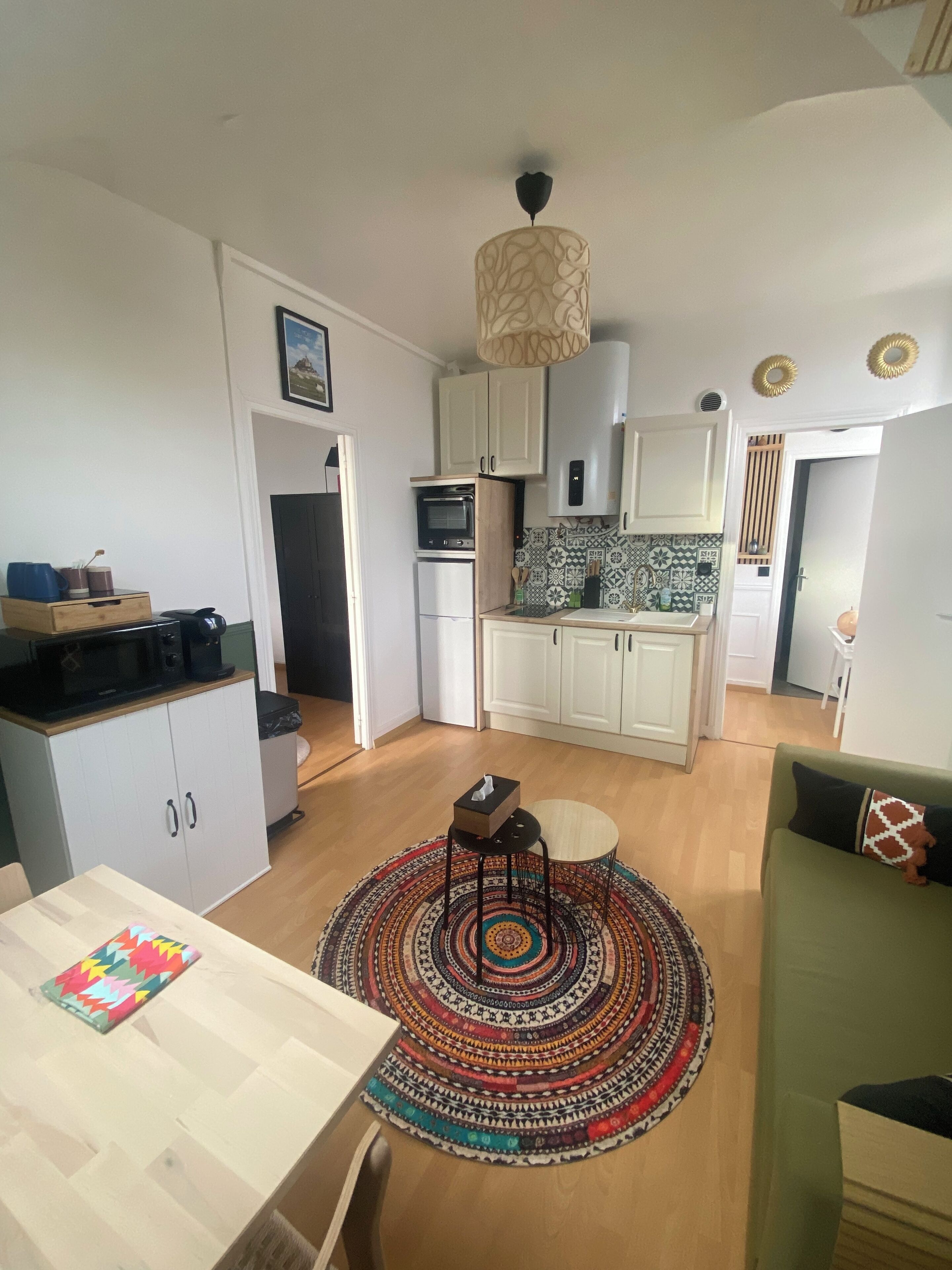 Cocina compacta con microondas, horno y armarios blancos. Cuenta con un comedor con mesa y sillas, y una alfombra colorida.