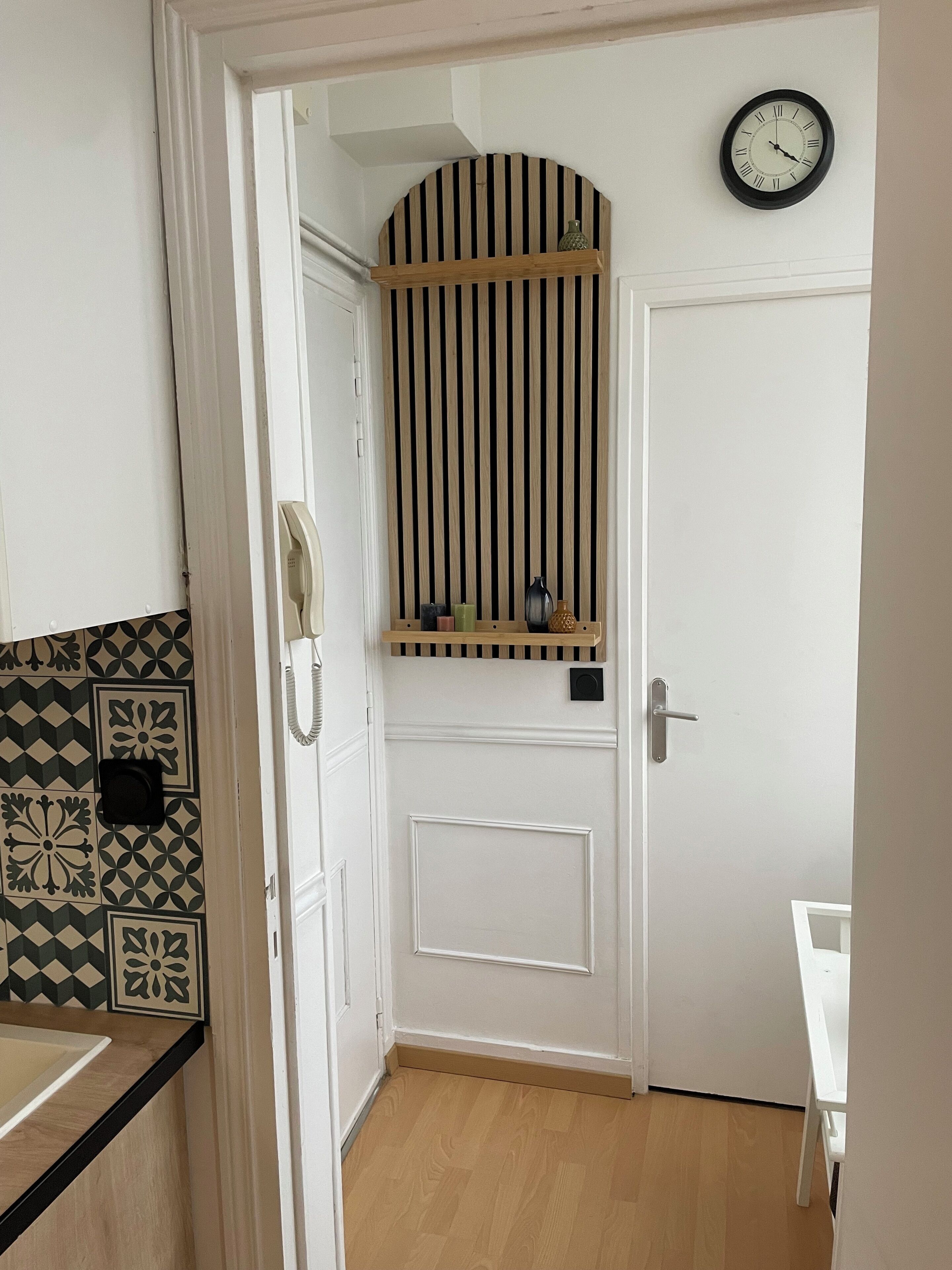 A hallway with a white door, a clock on the wall, and a wooden shelf with decorative items.