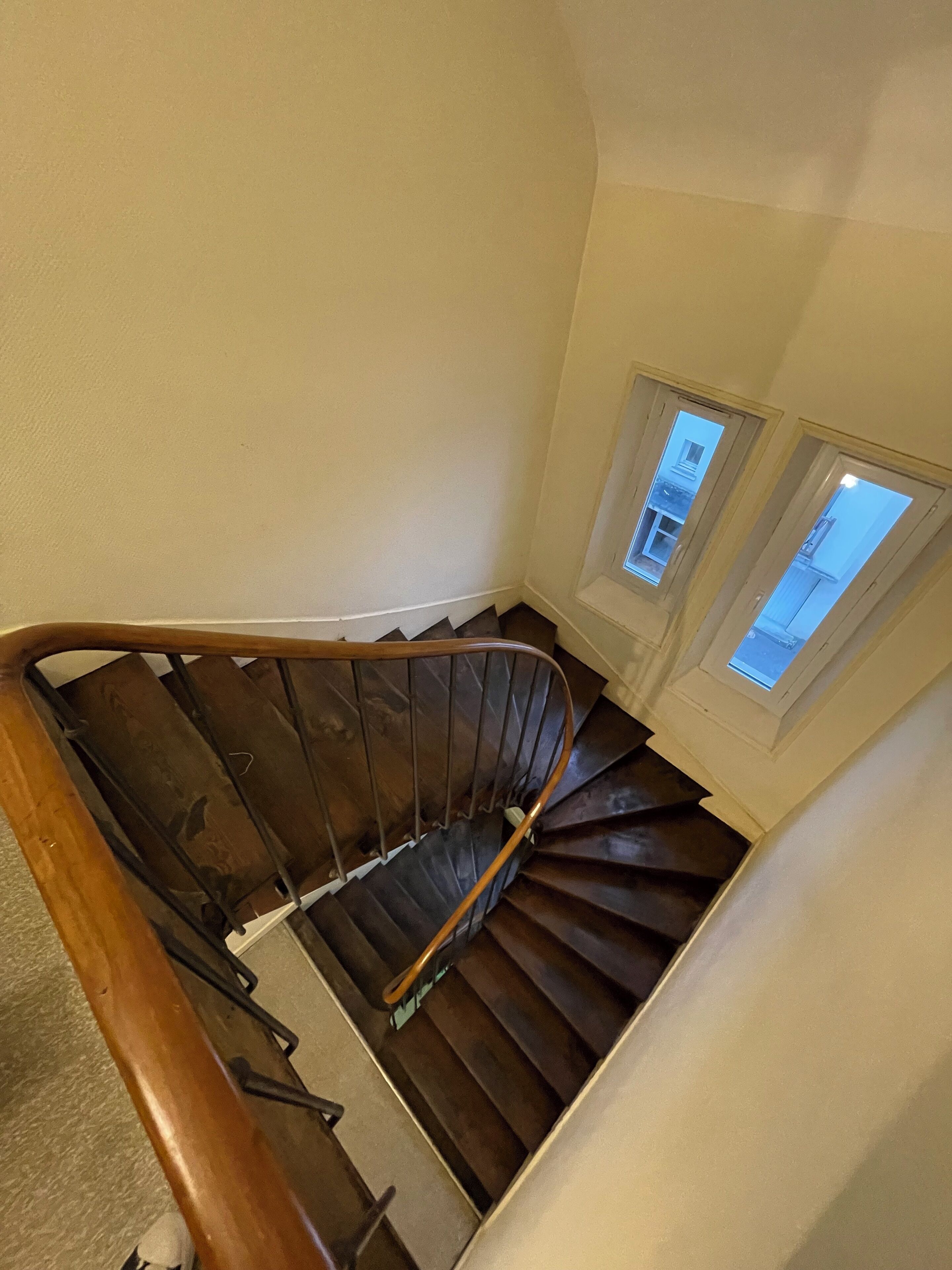 Un escalier en colimaçon avec des rampes en bois et des marches sombres, menant à un palier doté de deux fenêtres.