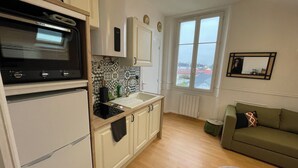 A compact kitchen with a microwave, sink, and cabinets. There's a window with a view of buildings and a green sofa in the living area. - A cocoon in the presbytery (with private parking)  (Caen)