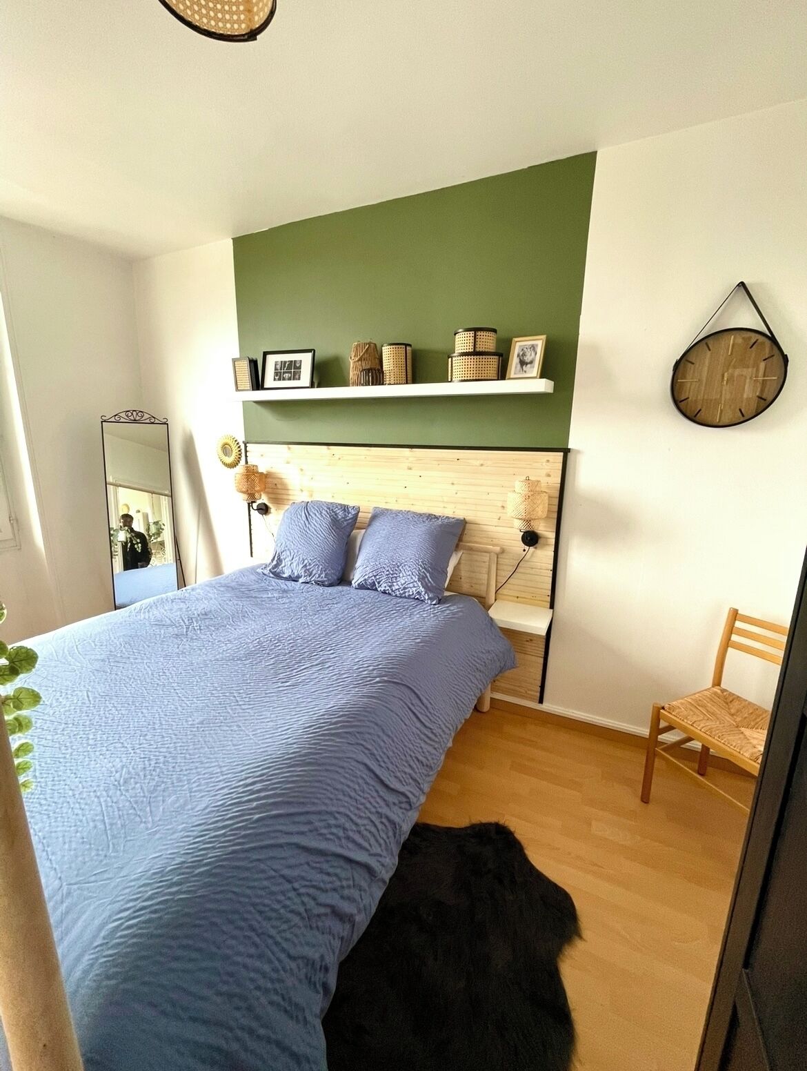 Quarto com uma cama grande, cabeceira de madeira, parede verde decorativa, uma cadeira de madeira e um relógio de parede redondo.