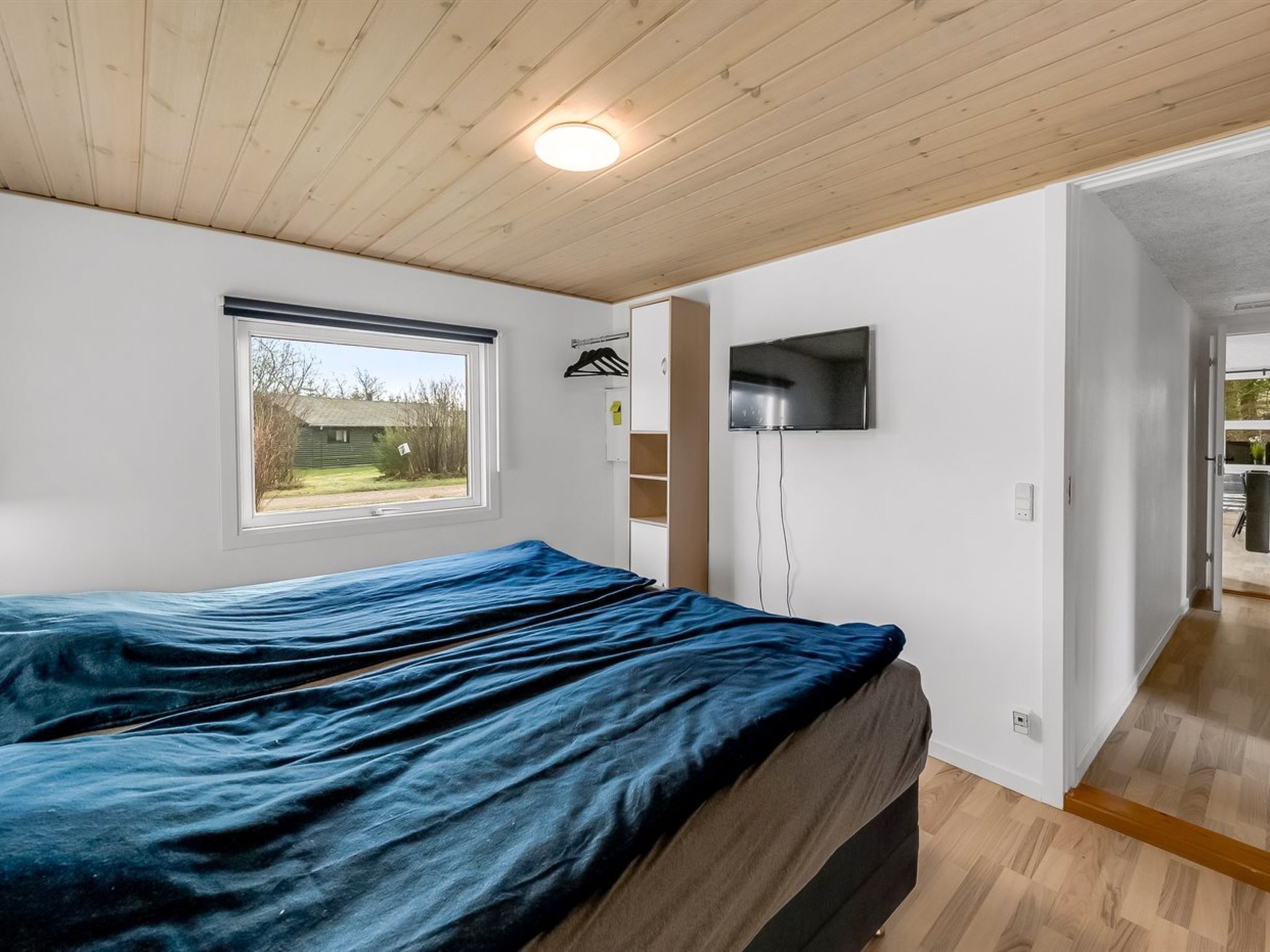 A bedroom with a bed, a TV mounted on the wall, a window with a view of the outside, and a door leading to another room.