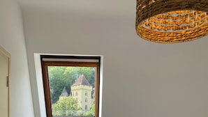 A bedroom with a bed, a small stool, a window with a view of a building, and a woven light fixture.