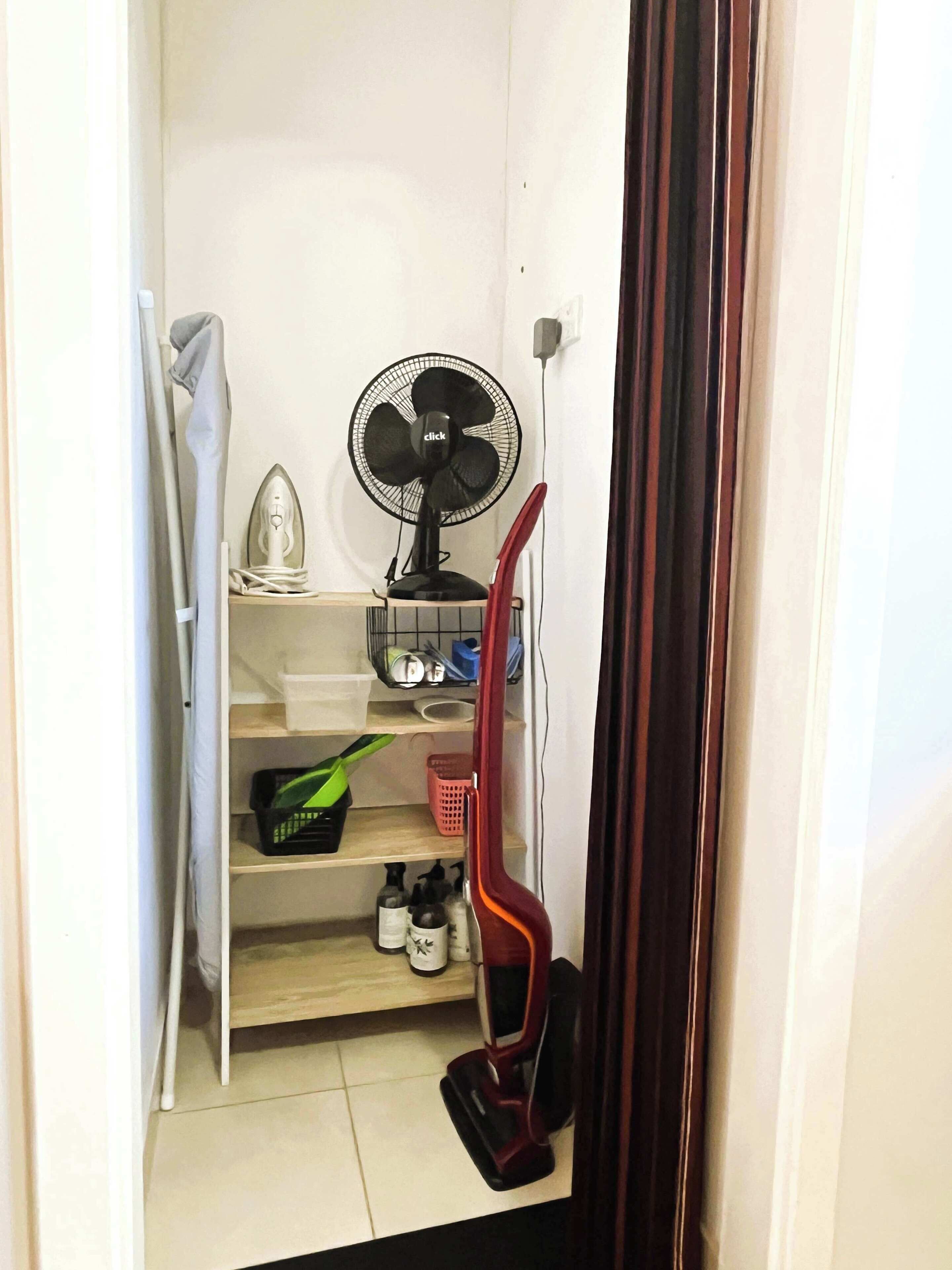 A narrow room with a fan, ironing board, shelves with storage bins, and cleaning supplies.