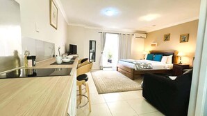 A modern hotel room with a kitchenette, a bed with a blue headboard, a black sofa, and a dining area with a table and chairs. - 45 Garden Guest Suite Studio (Bassendean)